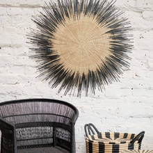 Load image into Gallery viewer, One Usiku Woven Wall Art pieces with a natural enter and black indigo edging displayed on a wall with a chair and side basket.
